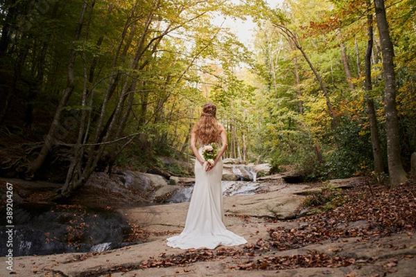 Fototapeta High angle rear view of beautiful young bride holding a bouquet on her back in the forest.