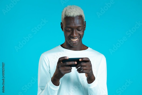 Fototapeta Handsome african man playing game on smartphone on blue studio wall. Using modern technology - apps, social networks.