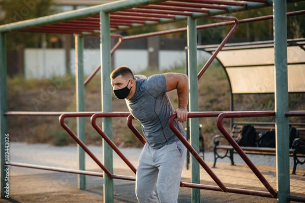 Fototapeta A young man does push-UPS, pull-UPS on a sports field in a mask during a pandemic at sunset. Sports, healthy lifestyle