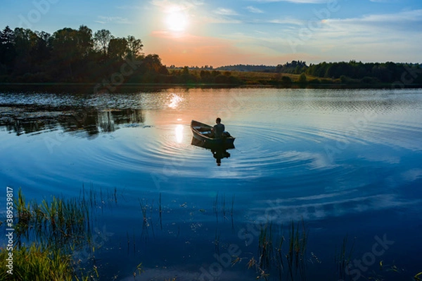 Obraz Mature caucasian man fishing in a small boat with a motor at sunset on the lake