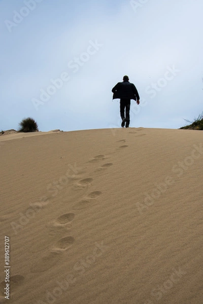 Fototapeta jeune homme marchant de dos de manière décidé sur la crête d'une dune de sable avec ses empreintes de pas visibles dans le sable