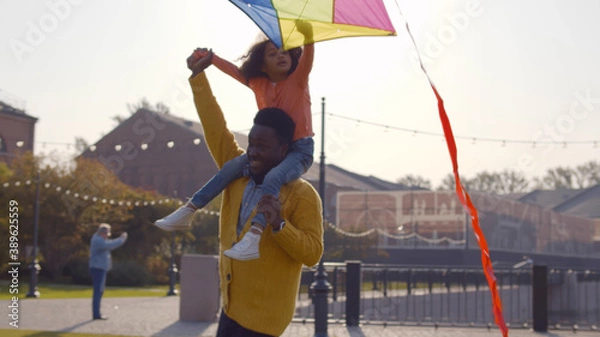 Fototapeta Cute african little girl sitting on shoulders of dad and playing with colorful kite
