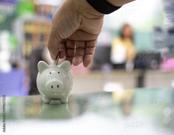 Obraz Close up. Hand of a man holding a coin for putting to white piggy bank in the bank office. Money-saving concept investment budget wealth business. Copy space on design, text. Bokeh, blurred background