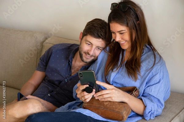 Fototapeta Young couple watching movie on cell phone and smiling on couch at living room