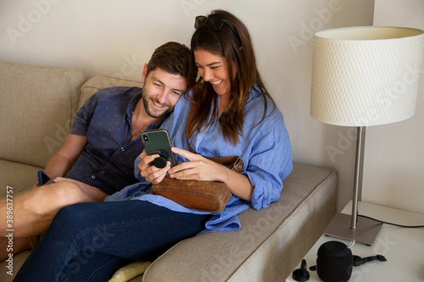 Fototapeta Young couple watching movie on cell phone and smiling on couch at living room