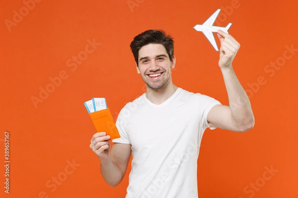 Obraz Smiling funny young traveler tourist man 20s wearing white t-shirt hold passport ticket air plane isolated on orange background. Passenger traveling on weekend getaway. Air flight journey concept.