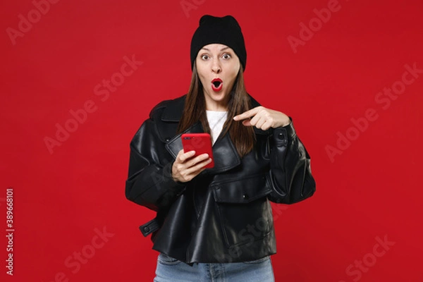 Fototapeta Shocked amazed young brunette woman 20s wearing casual black leather jacket white t-shirt hat pointing index finger on mobile cell phone typing sms message isolated on red background studio portrait.