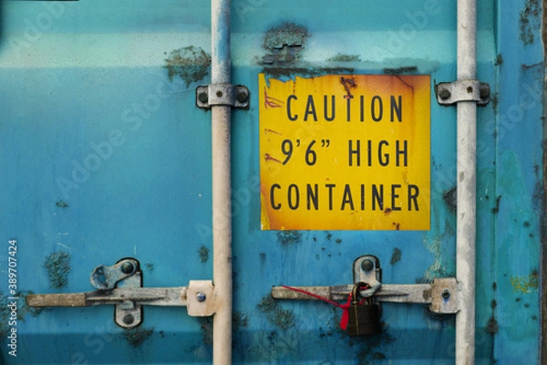 Obraz The back door of blue heavy-duty container with locks and sealed padlock. Container is very rusty and aged. Yellow warning sign about high container height is marked on the door