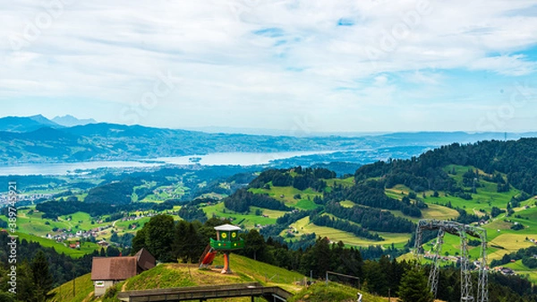 Fototapeta Paysage de montagne avec une vallée verdoyante,  des jeux, des arbres et de la forêt et des montagnes avec des cimes enneigées à l'arrière plan dans un ciel nuageux.