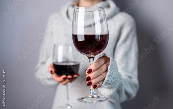 Fototapeta A woman in a white sweater is holding two glasses of red wine and holding out one of them. Close up