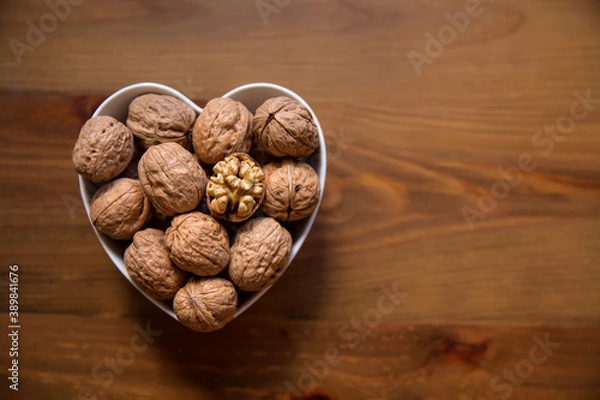 Obraz Whole walnuts and one cracked broken heart in a white heart-shaped bowl on a wooden background. Top view. Place for text.