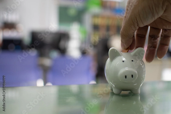Obraz Close up. Hand of a man holding a coin for putting to white piggy bank in the bank office. Money-saving concept investment budget wealth business. Copy space on design, text. Bokeh, blurred background