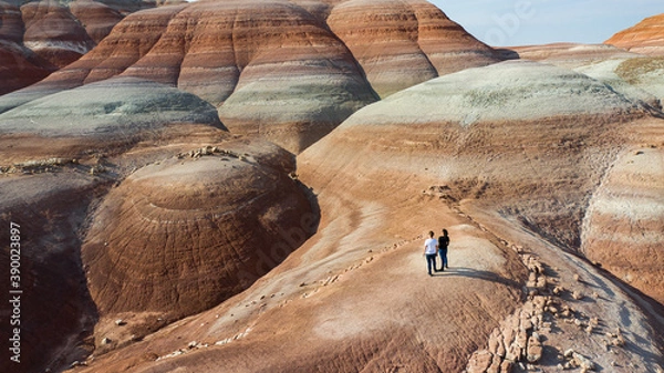 Fototapeta Couple looking out into a desert landscape resembling planet Mars. Rock layers with orange, red, blue, purple, and orange hues in the sediment can be seen 