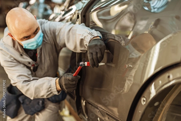 Fototapeta A car factory worker wearing a protective medical mask removes a metal defect with a hand tool.