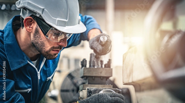 Obraz Male engineer in blue jumpsuit and white hard hat operating lathe machine.	
