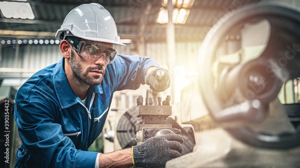 Obraz Mężczyzna inżynier w niebieskim kombinezonie i białym kasku obsługującym tokarkę.