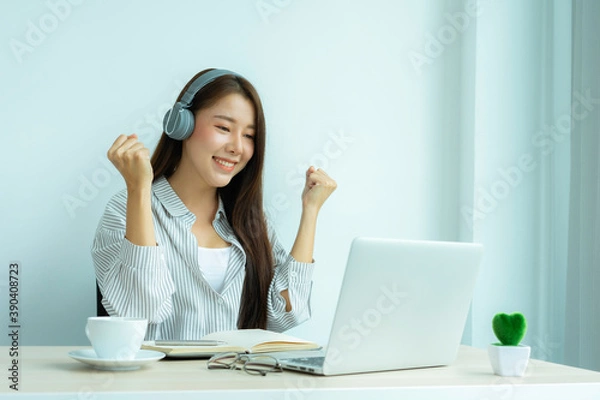 Fototapeta A young Asian woman who looks happily wears headphones. Watch webinars, listen to online courses, communicate by video conferences and learn languages, laughing, studying with teacher lessons via vide