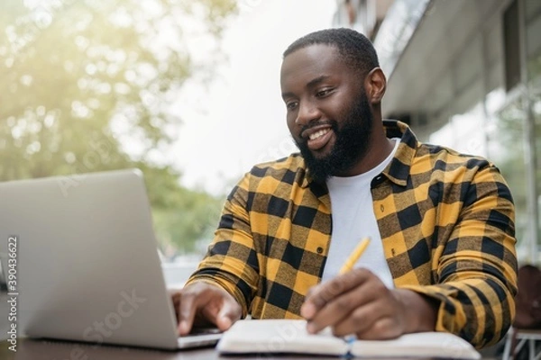 Fototapeta Young handsome African American man using laptop computer, taking notes, planning start up, working online. Portrait of happy student studying, learning languages, online education concept