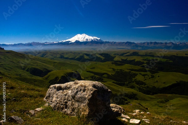 Fototapeta An excellent view from the plateau to the extinct volcano and the snowy peak of Mount Elbrus. Below is a green gorge. Stone in the foreground