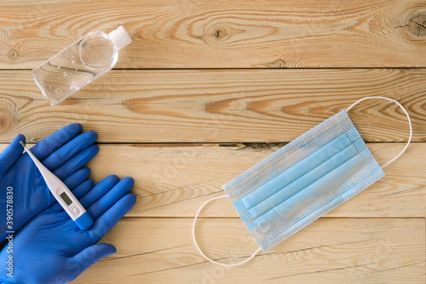 Fototapeta two hands in blue gloves hold a white thermometer, a blue mask and an antiseptic in a transparent plastic bottle on a wooden background