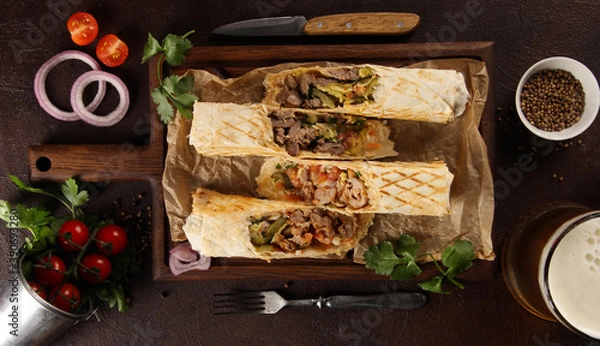 Fototapeta Shawarma with meat and vegetables. Flatbread with lamb, chicken, pickles, onions, mustard and sauce on a wooden board, beer and spices. Fork, knife, herbs, cherry tomatoes. Background image, top view