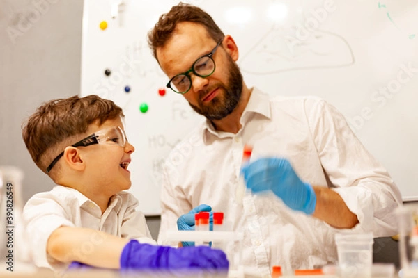 Obraz dad and son the child stayed at home conduct experiments on a white table, behind them on the wall is a white Board magnetic-marker