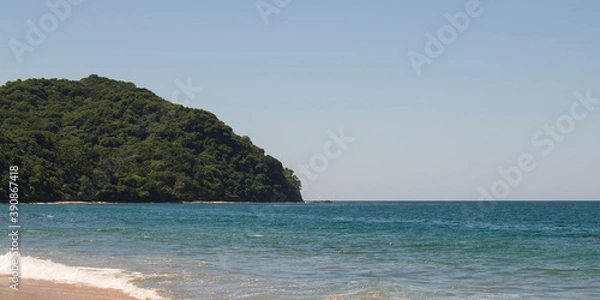 Fototapeta Beautiful landscape of a beach in the mexican pacific coast in a sunny day with a blue and clear sky, trees, palms and green hills, some waves and sand in the beach