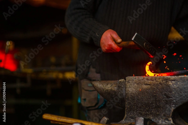 Fototapeta A close-up of the hands of a forge with a hammer striking a red-hot metal workpiece lying on an anvil and burning sparks scatter around