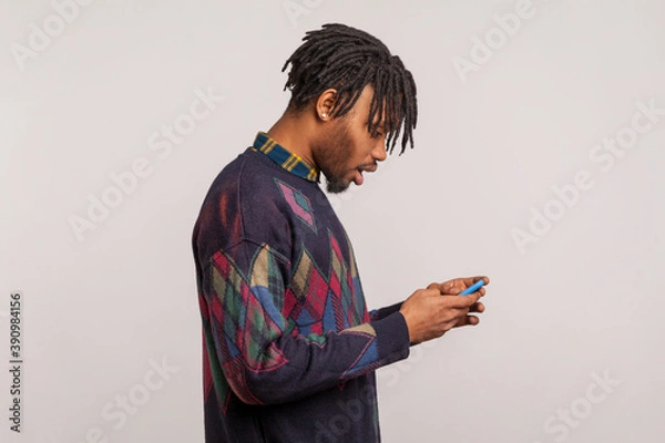 Fototapeta Extremely surprised african man with dreadlocks widely open his mouth holding mobile phone, wondered shocked with bonuses. Indoor studio shot isolated on gray background