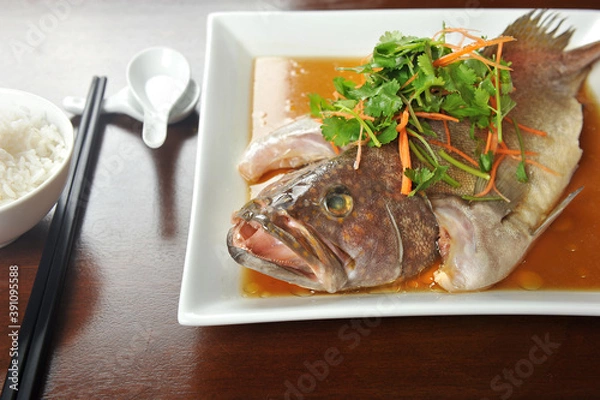 Fototapeta Steamed whole Grouper Fish in Soy Sauce of Chinese cooking style and garnish with shredded ginger and parsley, served on wooden table.
