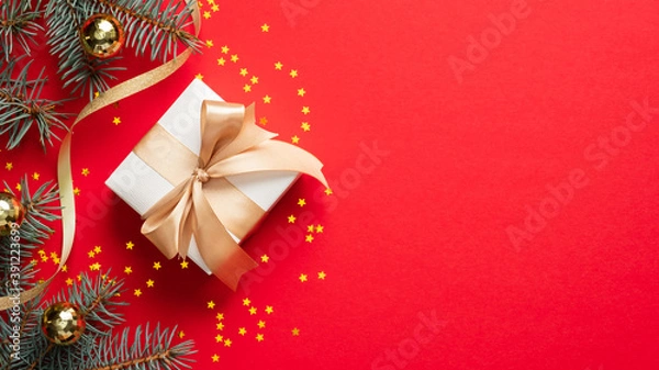 Fototapeta White Christmas gift box with golden ribbon bow, fir tree branches, balls, confetti stars on red background. Christmas present. Greeting card template, banner mockup for winter holidays.