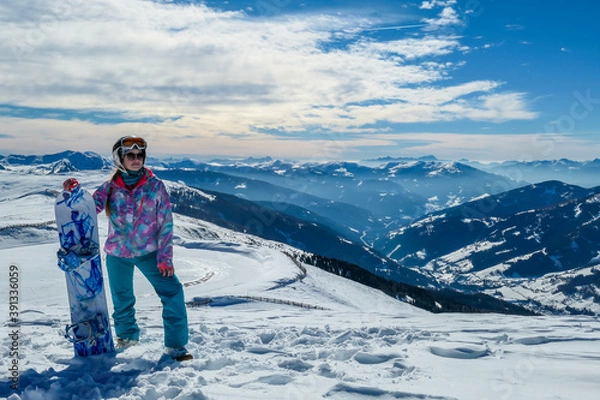 Fototapeta A woman posing on powder snow with her snowboard on top of Katschberg in Austria. Panoramic view on the surrounding mountains. Winter wonderland. Sunny winter day. She is full of energy and happy