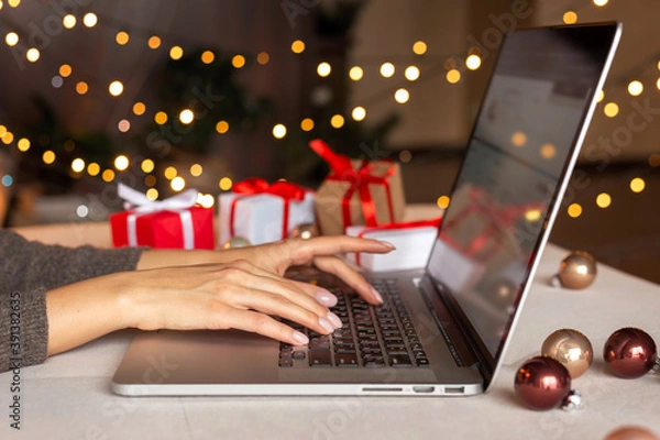 Fototapeta Christmas shopping online, sales and discounts promotions during winter holidays, online shopping at home and lockdown coronavirus. Female hands on the laptop with gifts and blurred bokeh lights