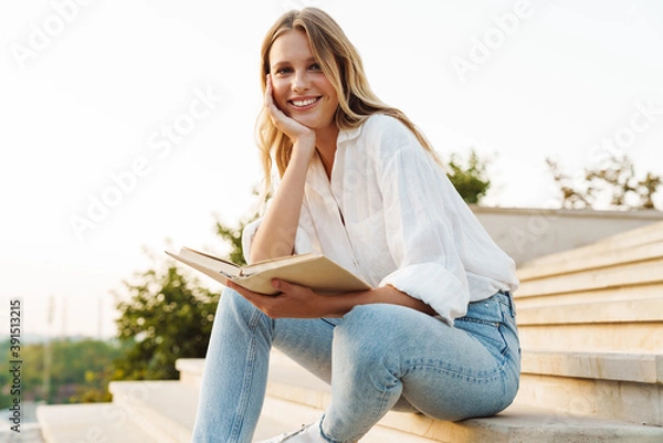 Fototapeta Beautiful happy woman smiling and reading book while sitting on staircase