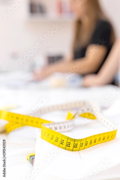 Fototapeta Close up of yellow tailor tape laying on the table with papers in sewing workshop with blurred tailor on background. Sewing accessories, tools. Measuring, centimeter