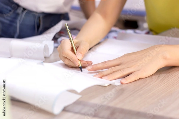 Fototapeta Fashion designer's hands. Closeup of hands drafting a new sewing pattern on paper, tailor working with a pencil, ruler, fabric, tool. Textile industry, hobby, workspace. Creation process. Drawing