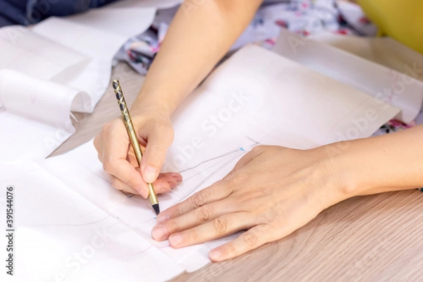 Fototapeta Fashion designer's hands. Closeup of hands drafting a new sewing pattern on paper, tailor working with a pencil, ruler, fabric, tool. Textile industry, hobby, workspace. Creation process. Drawing