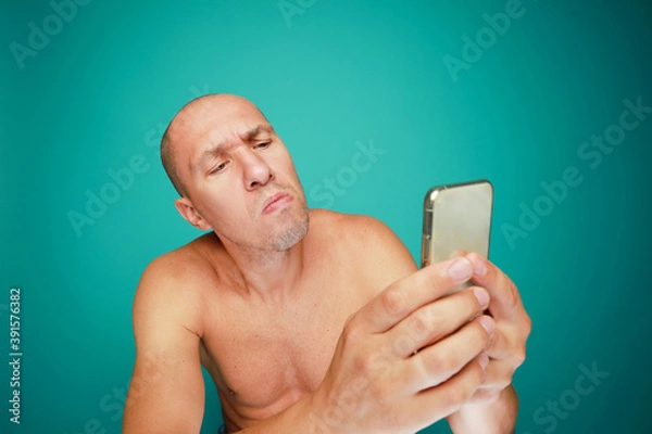 Obraz A funny bald and naked man with emotions holds mobile phone isolated on green background