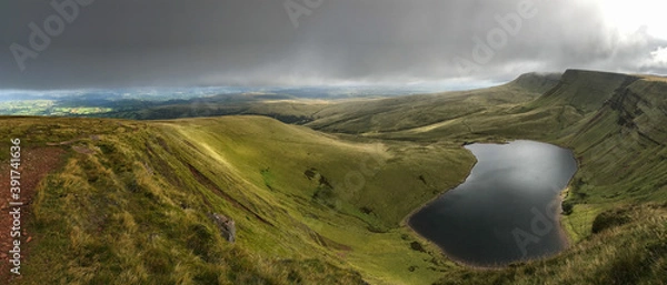 Fototapeta A panoramic view of the rolling green hills and mountains of the Brecon Beacons in Wales with a lake in the foreground and dark moody sky