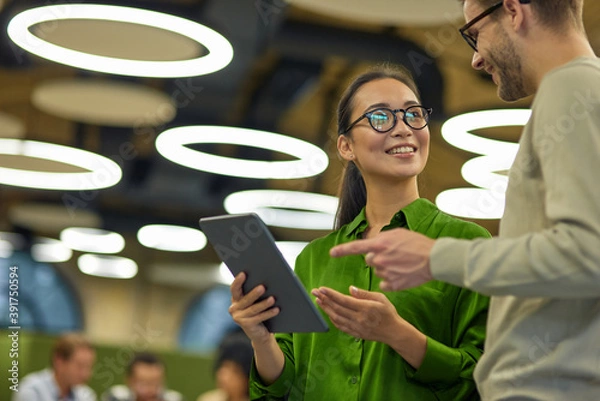 Obraz Discussing project. Young positive asian woman showing something on digital tablet to her male colleague while standing together in the coworking space or office