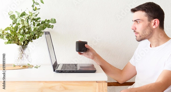 Obraz Retrato de un hombre adulto y apuesto caucásico sentado frente a su laptop y bebiendo una taza de café mientras la usa en un apartamento brillante y blanco con luz natural.