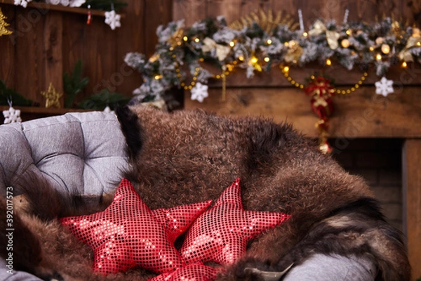 Fototapeta The comfort of Christmas in front of the fireplace arm chair with leather in dark colors. fur skin lying on a rattan chair. red pillows with shiny elements are sewn in the shape of a star