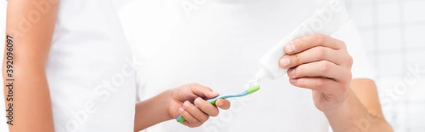 Fototapeta Cropped view of child holding toothbrush while standing near man squeezing toothpaste, banner