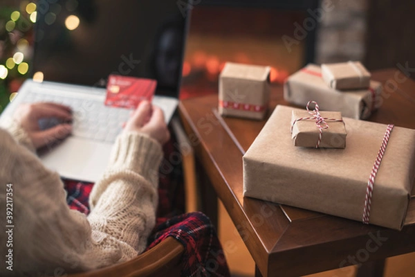 Fototapeta Christmas. Woman in sweater holding credit card using laptop for making order sitting at table with packaging gift near fireplace and christmas tree. Online shopping concept