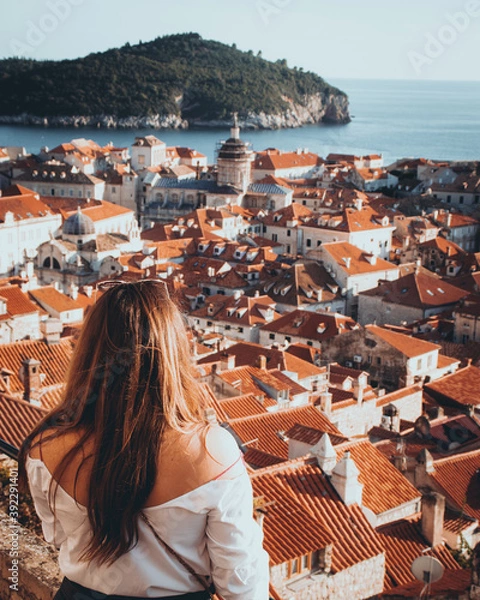 Fototapeta Vertical rear view shot of a girl standing on the city walls of Dubrovnik, looking towards the old red rooftops and the churches in the tightly packed city. Adriatic sea and islands in the distance