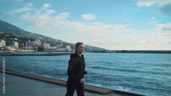 Fototapeta A young blonde girl in the early morning is engaged in jogging along the sea against the background of the lighthouse.