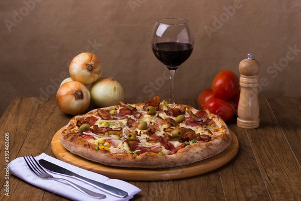 Fototapeta Portuguese Pizza made with ham, pea egg, heart of palm, pepperoni, onion and mozzarella and bacon. Served on a wooden board with red wine to accompany the food. Fork and knife.