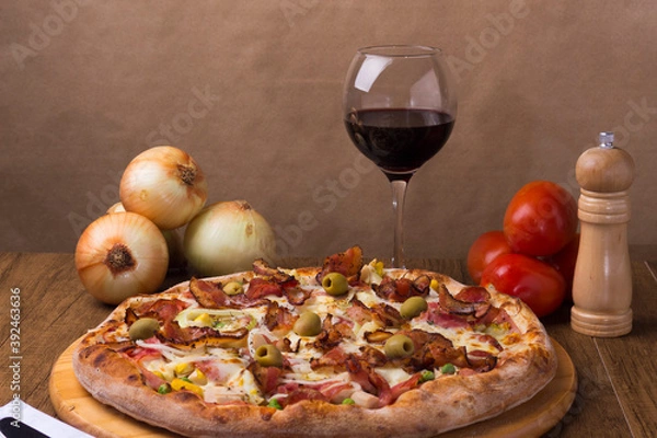 Fototapeta Portuguese pizza made with ham, pea egg, heart of palm, pepperoni, onion and mozzarella and bacon. Served on a wooden board with red wine to accompany the food. Centralized elements.