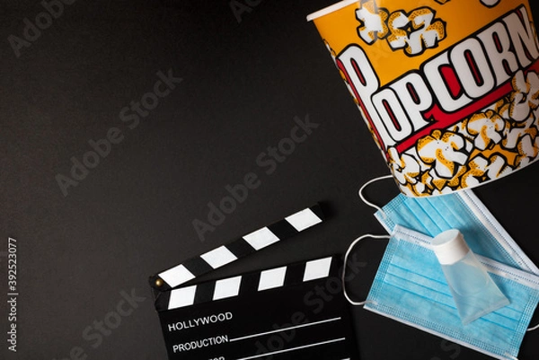 Fototapeta Movie clapperboard, hydrogel, face mask, and popcorn box on a black background during the second wave of the coronavirus pandemic. Covid concept.