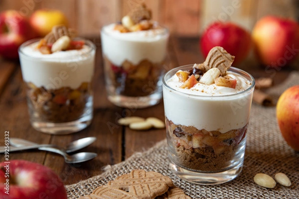 Fototapeta Kuchen im Glas mit Stücken vom Bratapfel, Zimt, Spekulatius, Apfel, Mandeln und Rosinen mit einer Creme aus Quark oder Mascarpone auf einem Tisch aus Holz in einer gemütlichen Küche zu Weihnachten